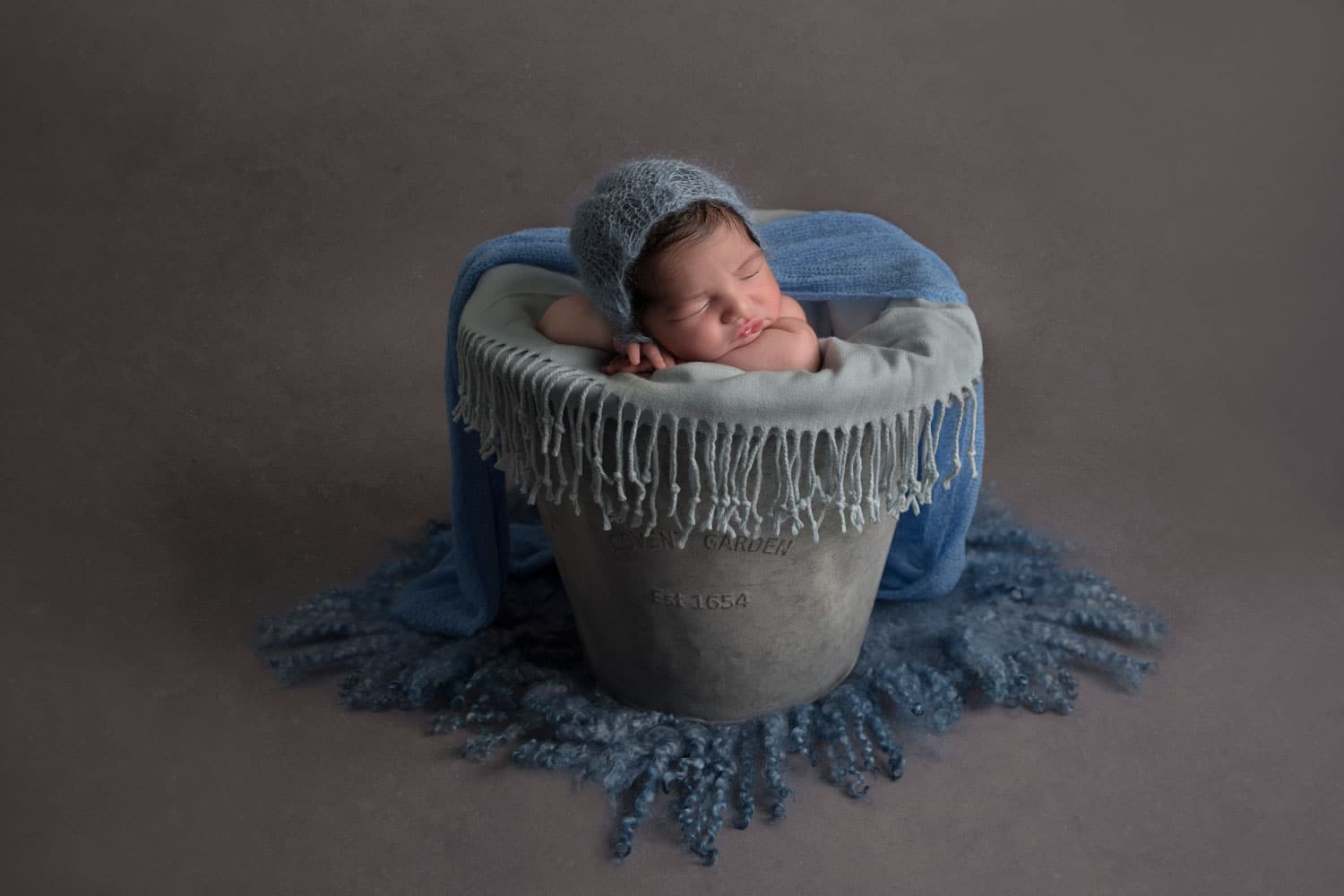 little newborn boy sitting in grey basket with blue hat and blanket
