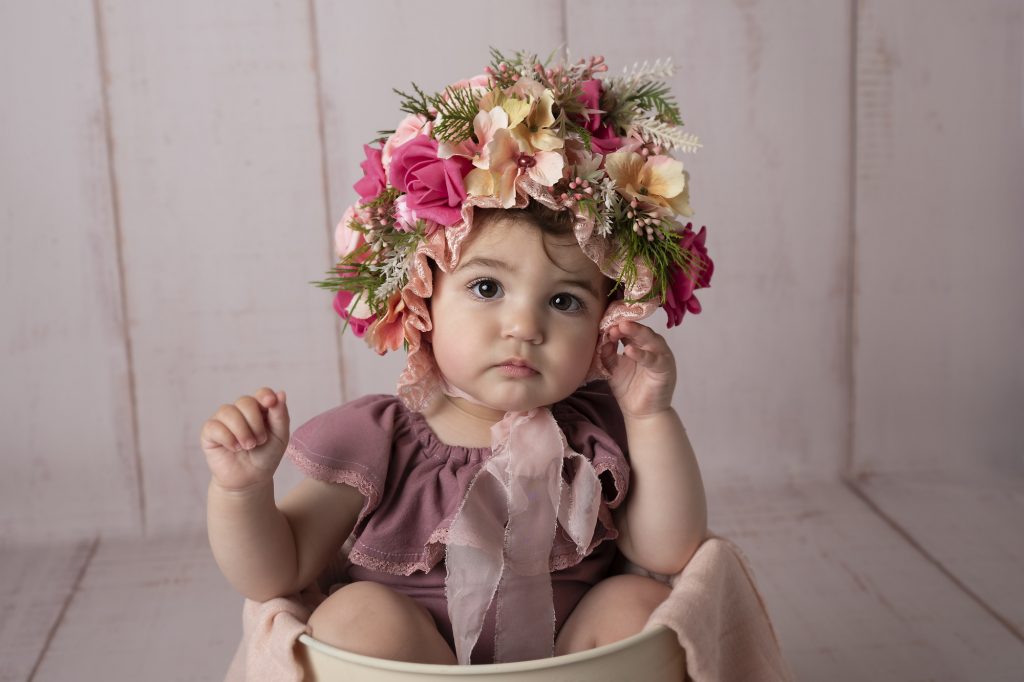 baby girl is sitting in bucket and wearing flower bonnet photographed by Baby Photographer Manchester