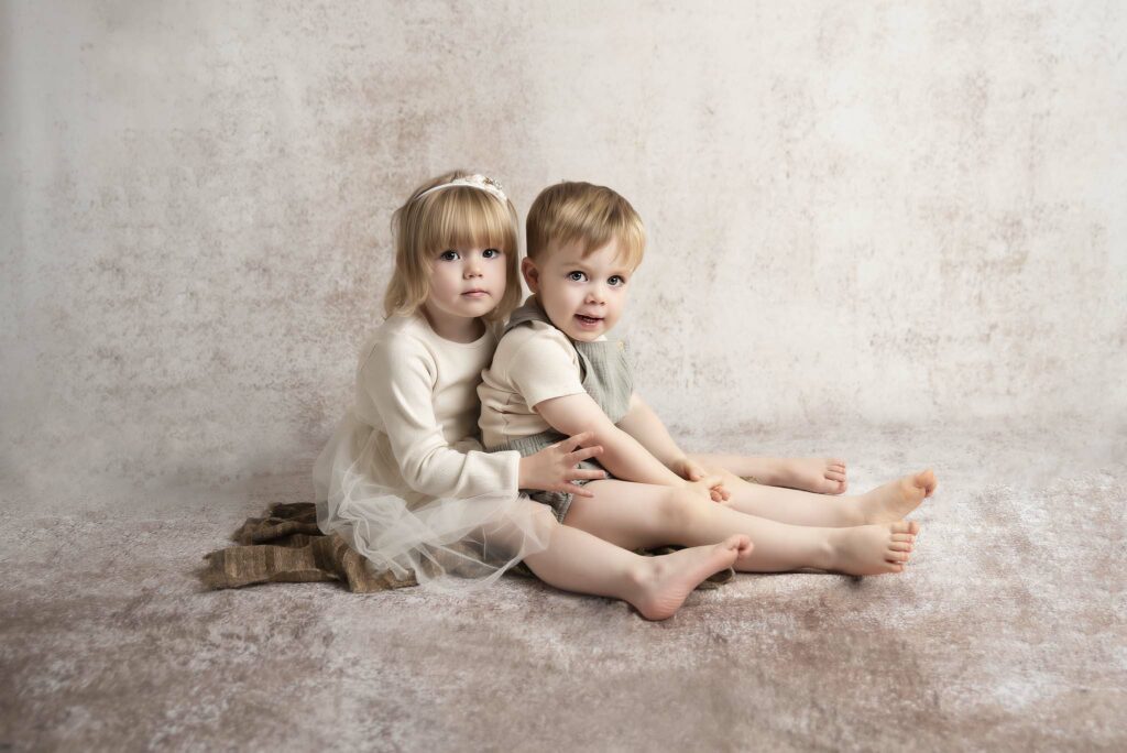 sitter session with siblings sitting and hugging each other by a creamy background in a baby photography studio in manchester