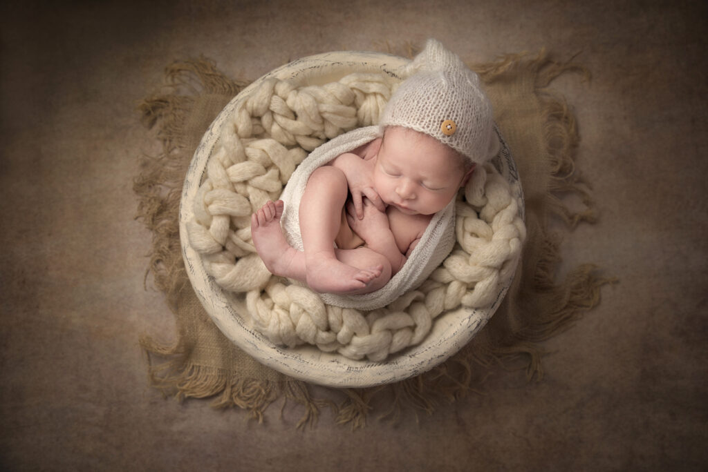 newborn baby in a white bowl and brown backdrop wearing a creamy hat in a cozy pose