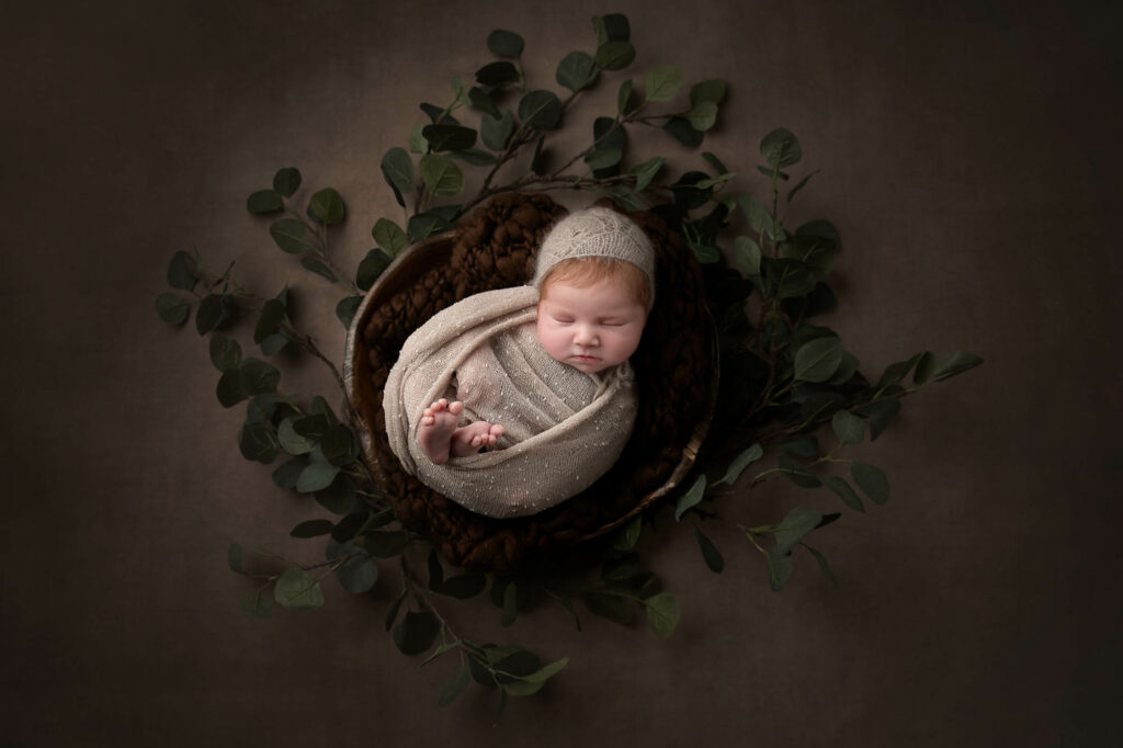 newborn baby sleeping on a basket with creamy wrap and leaves around him taken by newborn photographer Dora Horvath